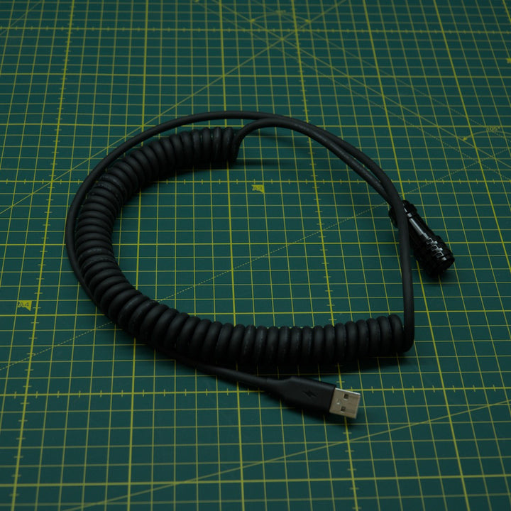 A coiled black USB cable with a USB-A connector on one end and a smaller black connector on the other, placed on a green cutting mat with yellow grid lines.