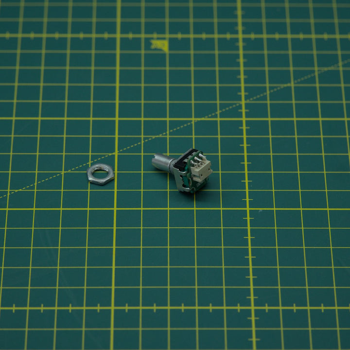 A small metal hardware component with a hex nut beside it, placed on a green cutting mat with yellow grid lines.