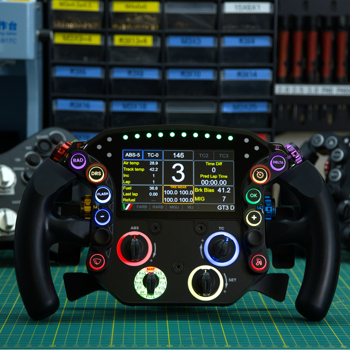 A high-end racing steering wheel with an integrated digital display and illuminated control buttons, set against a blurred workshop background.