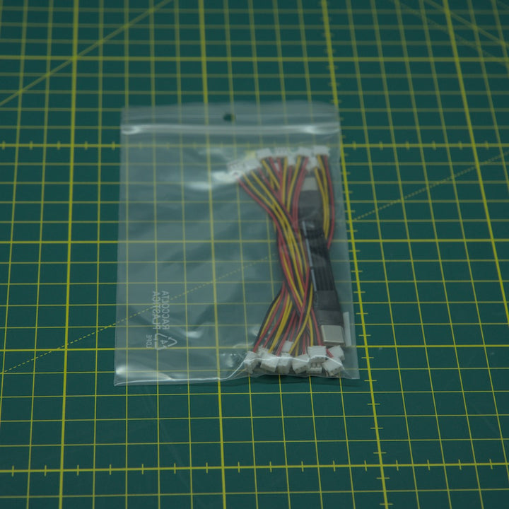 A small clear plastic bag containing a multi-pin ribbon cable with white connector ends, placed on a green cutting mat with yellow grid lines.