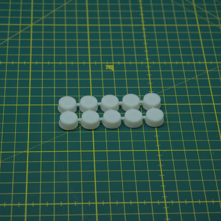 Eight small white round plastic pieces arranged in two rows on a green cutting mat with yellow grid lines.