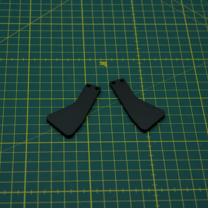 Two small black metal components with pre-drilled holes, placed side by side on a green cutting mat with yellow grid lines.