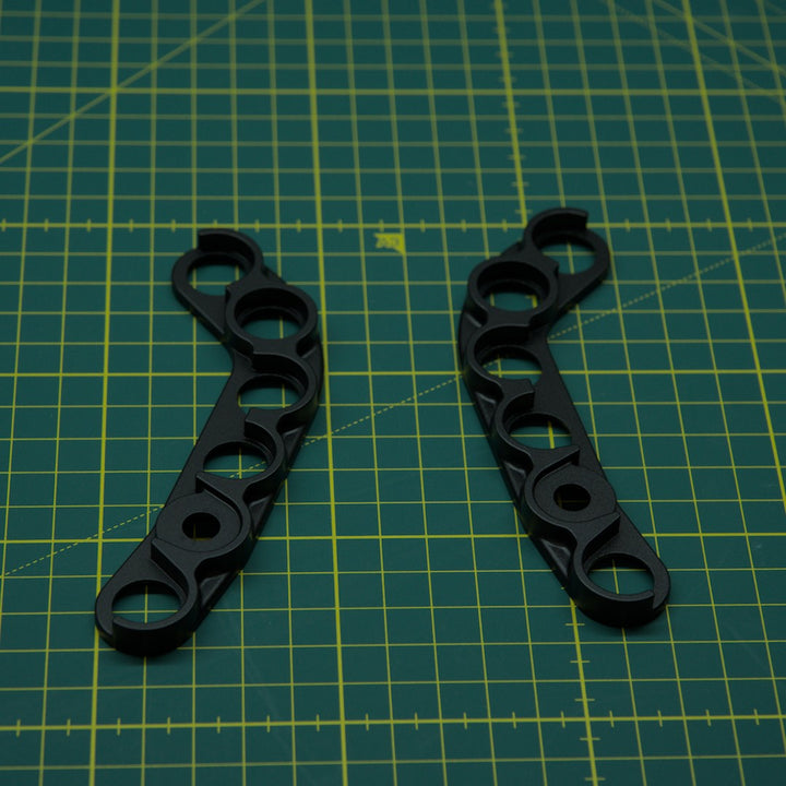 Two black metal mechanical brackets with circular cutouts, displayed on a green cutting mat with yellow grid lines.