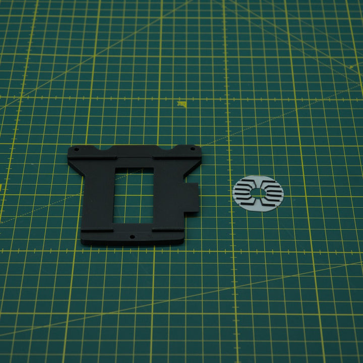A black plastic mounting bracket and a small circular white and black sticker are placed on a green cutting mat with yellow grid lines.